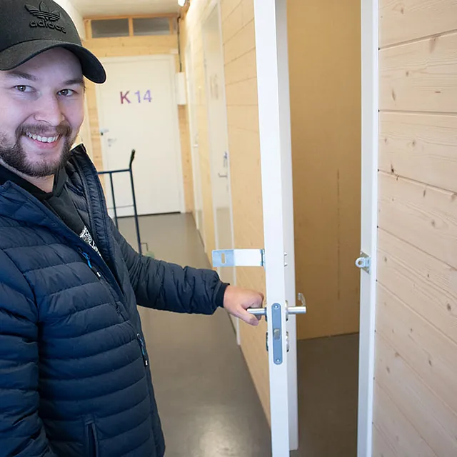 Smilende mann åpner dør til lagerrom i gang med lyse trefinérvegger og nummererte dører.