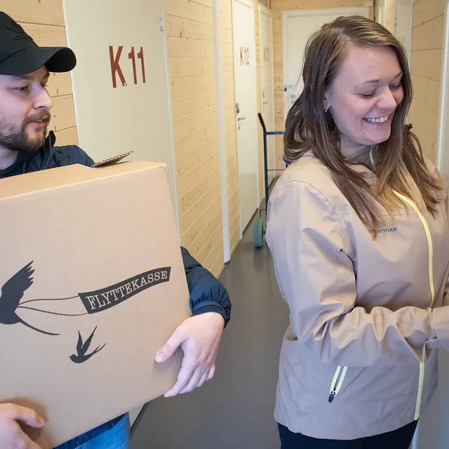Kvinne åpner dør til minilager mens mann holder flyttekasse i gang med lyse tredører merket med nummer.