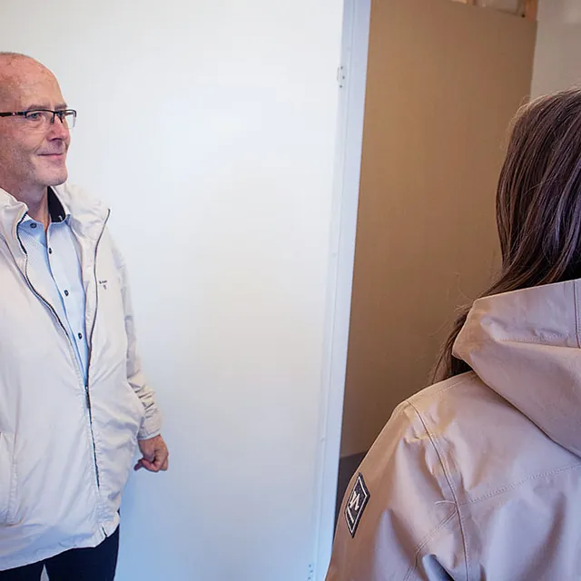 To personer står og snakker ved inngang til lagerrom med hvit dør og lyse vegger.