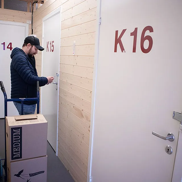 Mann låser opp dør til bod merket K15 i gang med flyttekasser og tralle.