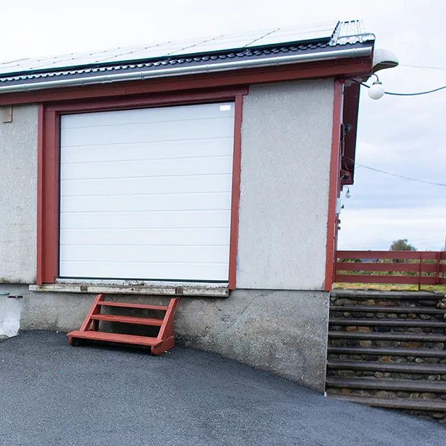 Grå lagerbygning med rød ramme og liten trapp, ved asfaltert plass og grønne omgivelser.